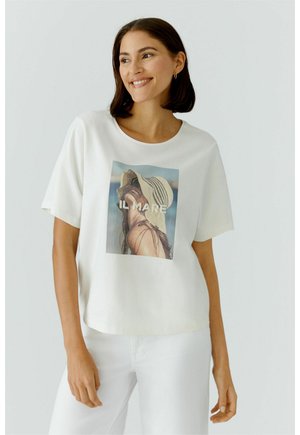 Junge Frau lächelt, trägt ein weißes T-Shirt mit einer Grafik einer Frau mit Sonnenhut und dem Text "IL MARE" auf der Vorderseite sowie weiße Hosen.