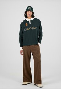 Polo verde de manga larga con cuello blanco y bordado "Social Club", combinado con pantalones anchos marrones y zapatos crema.