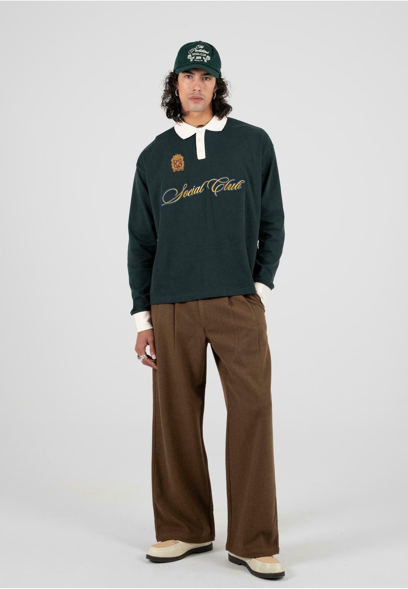 Polo verde de manga larga con cuello blanco y bordado "Social Club", combinado con pantalones anchos marrones y zapatos crema.