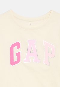 Κρεμ T-shirt για παιδιά με μεγάλες, έντονες γράμματα "GAP" σε αποχρώσεις του ροζ στο στήθος και στρογγυλή λαιμόκοψη.
