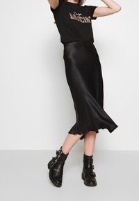 Falda deslizante de satén negro con dobladillo acampanado, combinada con una camiseta negra con el texto metálico "Keep Dancin'" y botas de tobillo negras con hebillas.