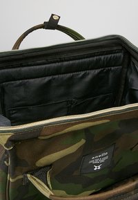 Reppuvaatekuvioinen laukku, jonka vetoketju on auki ja paljastaa pehmeän mustan sisustan sekä kankaasta valmistetun etiketin, jossa on valkoista tekstiä.