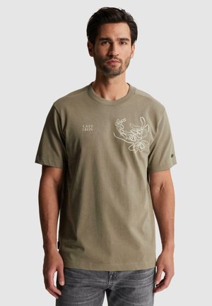 Homme portant un t-shirt vert olive avec un motif floral blanc et le texte "CAST IRON" sur la poitrine, associé à un jean gris foncé, debout devant un fond blanc.