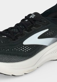 Scarpa da corsa bianca e nera con lacci neri, logo bianco sul lato e suola texturizzata su sfondo neutro.