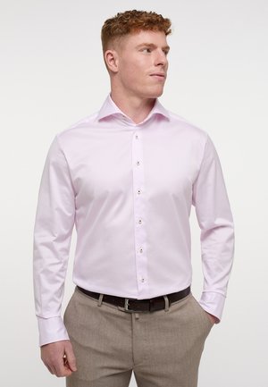 Helles pinkfarbenes Langarmhemd mit spitzem Kragen, Knopfleiste und subtiler glänzender Oberfläche. Getragen mit hellbeigen Hosen und einem dunklen Gürtel.