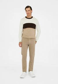 Man in beige broek en een kleurbloktrui met witte, donkerbruine en beige strepen, dragend witte sneakers, staande tegen een eenvoudige achtergrond.