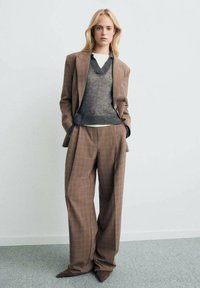 Costume à carreaux marron avec un pantalon oversized et un pull en maille gris superposé sur un haut blanc. Comprend des chaussures pointues et une coupe décontractée.