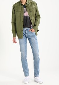 Camicia in twill di cotone verde con tasche frontali, indossata sopra una t-shirt grafica, abbinata a jeans azzurri chiari e sneakers bianche.