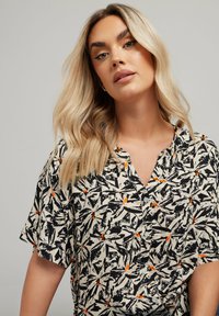 Blusa a maniche corte con colletto in crema con stampa di foglie nera e arancione. Presenta bottoni sul davanti e una vestibilità ampia.