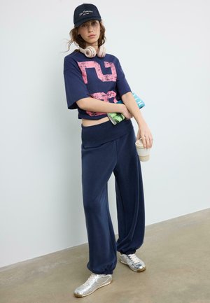 Mlada ženska v širokih mornarsko modrih hlačah, mornarsko modri kratki majici s rožnatim potiskom, z kapa, slušalkami, drži torbico in skodelico kave, stoji v notranjosti.