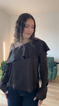 Czarny top z odsłoniętymi ramionami, z falbaną przy dekolcie i długimi rękawami, wykonany z gładkiej tkaniny, zestawiony z ciemnoniebieskimi dżinsami.