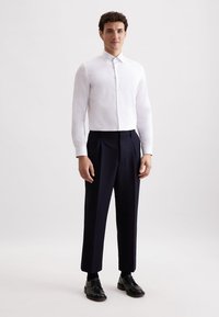 Weißes Hemd mit langen Ärmeln, kombiniert mit navyblauen, gefalteten Hosen und schwarzen Schnürschuhen vor einem neutralen Hintergrund.