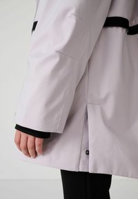Svetlý levanduľový nepremokavý kabát s jemným leskom, s čiernymi akcentmi, bočným rozparkom a priliehavým rukávom siahajúcim po zápästie.