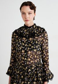 Kvinne med kort krøllete hår iført en gjennomsiktig svart blomstret kjole med rysjedekolt og perleknapper, mot en enkel hvit bakgrunn.