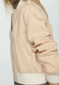 Světlobéžová bomber bunda s žebrovanými bílými manžetami, zapínáním na zip a bočními kapsami. Hladká tkanina a mírně přiléhavý střih.