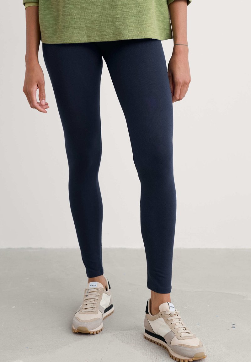 Persoon die marineblauwe legging draagt, beige en witte sneakers en een groene trui, staand op een lichtgrijze vloer tegen een effen muur.