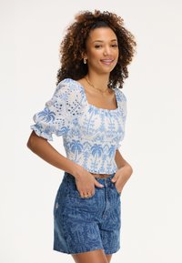 Top court à motifs bleus et blancs avec des manches bouffantes, corsage froncé, associé à un short en denim bleu taille haute avec des designs complexes.