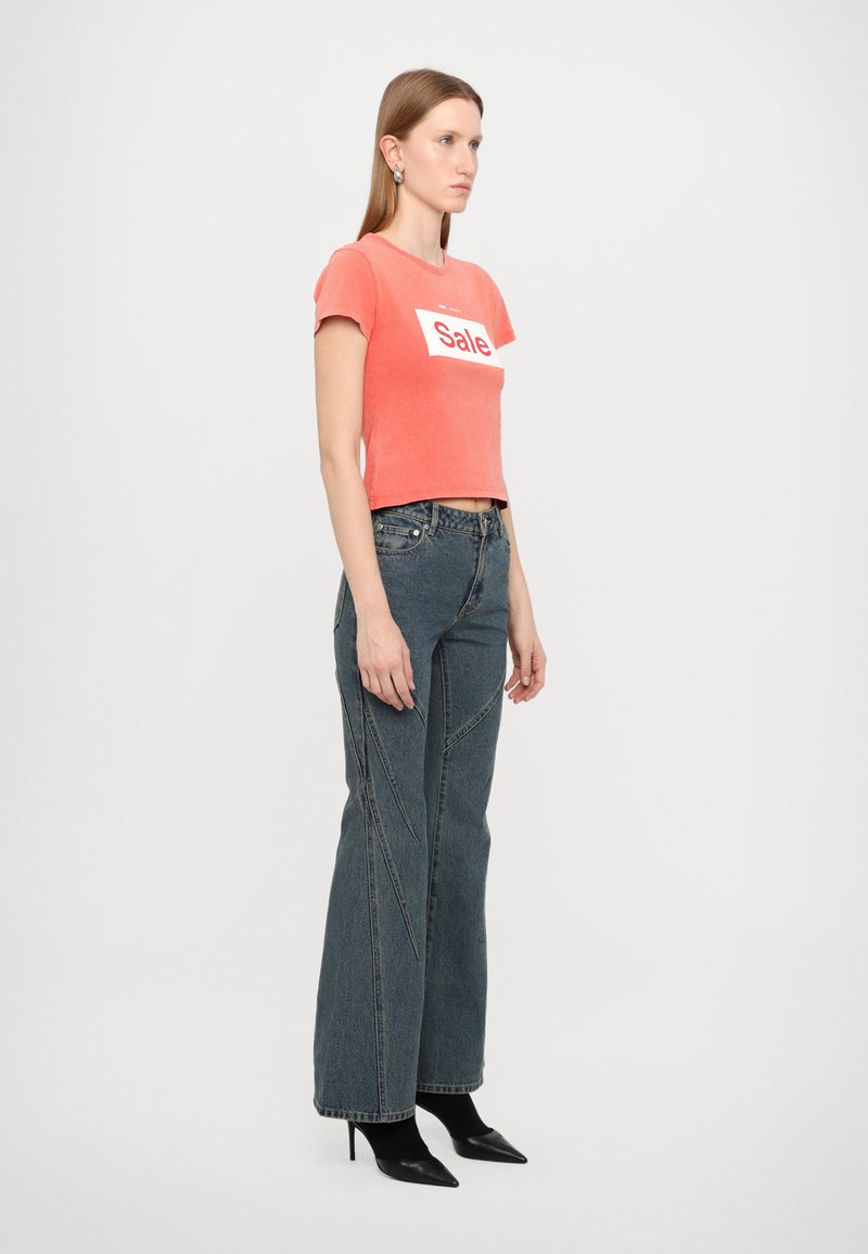 Camiseta corta coral con estampado blanco de "Rebaja", combinada con unos pantalones de mezclilla oscuros de pierna ancha y cintura alta que presentan detalles de costura y tacones negros.