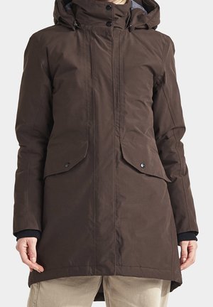 Brun lang vinterfrakke med hætte, frontlommer med klap og knaplukning, båret over beige bukser.