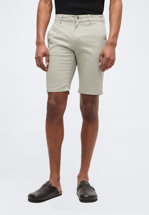 Homme portant un short beige au genou, une chemise noire et des chaussures noires sans lacets, debout devant un fond clair uni.