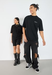 Two models wear black oversized t-shirts with a small logo. The male model's distressed jeans are paired with black sneakers; the female model wears black bike shorts and tall boots.