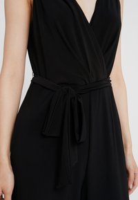 Mono negro con diseño envolvente, escote en V profundo y cinturón atado en la cintura. Confeccionado en una tela suave con una silueta ajustada y elegante.