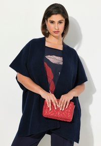 Marinblå stickad cardigan med korta ärmar, öppen front, texturerat tyg, kombinerad med en kviltad röd clutchväska. Enkel blommig mönstrad topp under.