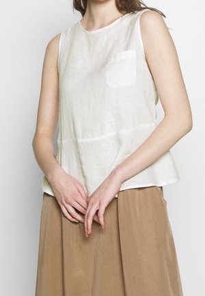 Ärmelloses weißes Leinenoberteil mit Fronttasche, getragen mit lockerem braunem Rock, die Hände sanft vor der Taille geschlossen.