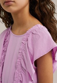 Orgona színű pamut blúz fodros díszítéssel a vállakon és az elején, rövid légies ujjakkal és puha textúrával.