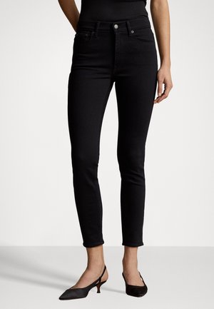 Femme portant un jean noir ajusté et des chaussures à talons petits noirs slingback, debout sur un fond clair uni.