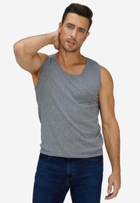 Uomo con capelli scuri che indossa una maglietta grigia senza maniche e jeans blu, che si tocca il collo con la mano sinistra.