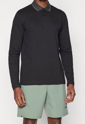 Homme portant un polo noir à manches longues avec un col gris et un short vert clair avec un petit logo sur la jambe gauche.