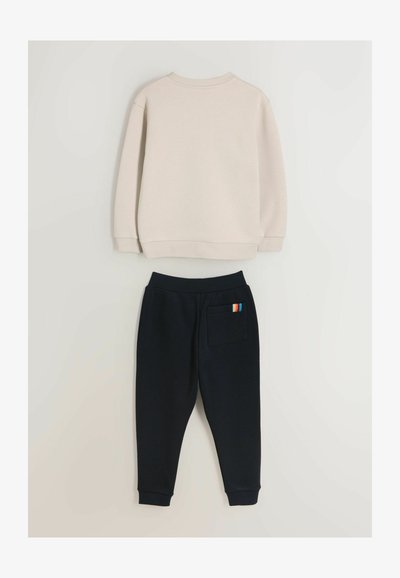 Bēšs džemperis ar garām piedurknēm un ribotām aprocēm, kopā ar tumši ziliem sporta biksēm ar daudzkrāsainu svītru kabatas detaļu.