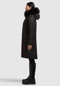 Musta toppatakki, jossa on karvareunainen huppu, pitkät hihat ja etutaskut. Päällä mustat polven korkeudelle ulottuvat saappaat ja leggingsit. Sivuprofiilinäkymä.