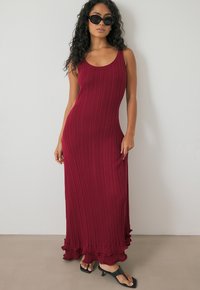 Rippent, hoch-elastisches Maxikleid in burgundy mit runden Ausschnitt, ausgestelltem Rüschensaum und glatter Textur, kombiniert mit schwarzen Sandalen und Sonnenbrille.
