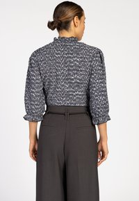 Vrouw met een knot die een hoogsluitend, patroon blouse met 3/4 mouwen en donkere, hooggerokte, wijde broek draagt, bekeken van achteren.