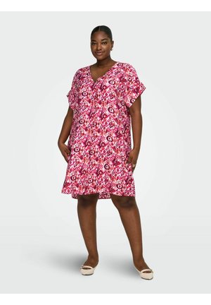 Kvinde stående iført en kortærmet kjole til knæet med abstrakte blomsterprint i pink, rød og hvid samt beige flade sko.