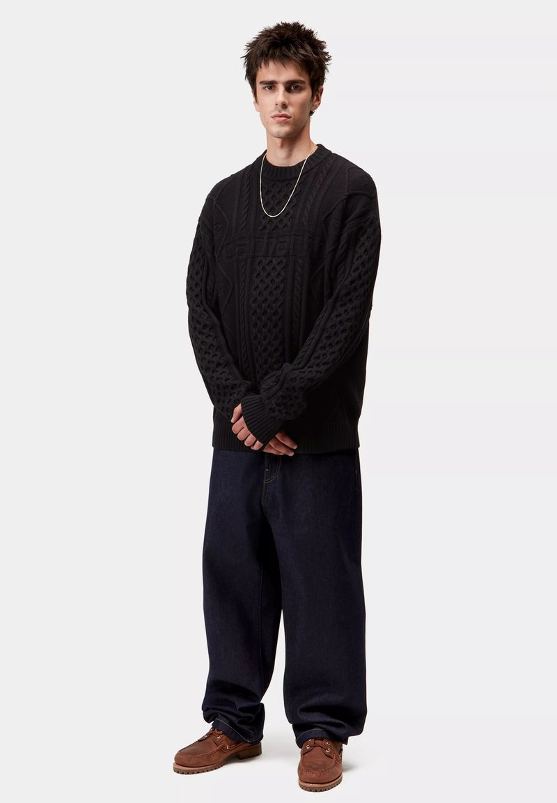 Suéter negro de punto con patrones texturizados, combinado con unos jeans de denim oscuros y sucios, y zapatos marrones con cordones. El modelo está de pie con los brazos cruzados.