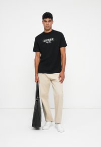 Camiseta negra con el logo "GUESS" en blanco, combinada con pantalones beige y zapatillas blancas. El modelo lleva una bolsa tote negra.