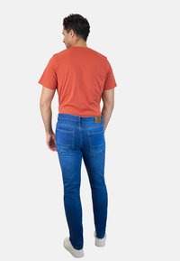 Camiseta naranja de hombre combinada con jeans ajustados azules. Los jeans tienen bolsillos traseros y un parche de cuero con el logo en la cintura. Zapatillas blancas.