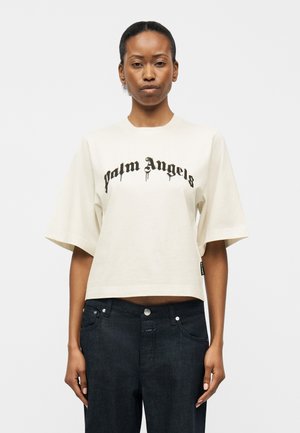 Γυναίκα που φοράει ένα κοντό T-shirt εκρού με την επιγραφή "Palm Angels" σε μαύρη γοτθική γραμματοσειρά, σε συνδυασμό με χαλαρά σκούρα τζιν.