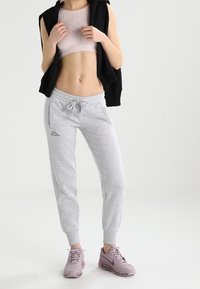 Graue Jogginghose mit elastischen Bündchen, ein schwarzes ärmelloses Hoodie und ein hellrosa Sportoberteil. Tragen mit helllila Sportschuhen.