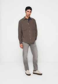 Chemise en velours côtelé marron avec deux poches sur la poitrine, associée à un jean gris clair et des sneakers crème. Le modèle se tient devant un fond blanc uni.