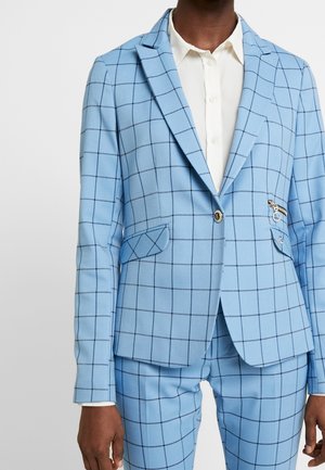 Person trägt einen hellblauen Anzug mit einem marineblauen Fensterkaro-Muster und einem weißen Hemd darunter.