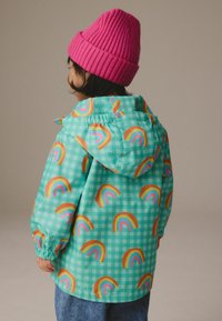 Veste de pluie colorée en turquoise avec un motif à carreaux ; présente des motifs de rainbow répétés dans des couleurs vives ; inclut une capuche et des poignets élastiques.