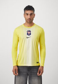 Dzeltena sporta krekls ar garām piedurknēm un horizontālu svītru rakstu, ar zilu emblēmu un melno Nike logo priekšā.