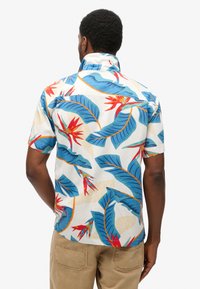 Camisa de manga corta en tejido blanco con un colorido estampado floral tropical que presenta hojas azules y flores rojas, cuello texturizado y corte relajado.