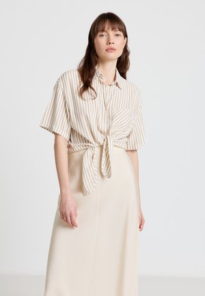 Frau trägt beige-weiß gestreiftes Kurzarmhemd mit vertikalen Streifen, das in der Taille gebunden ist, dazu cremefarbener, hoch taillierter Rock, steht vor einfarbigem Hintergrund.