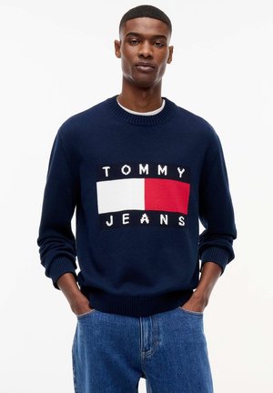 Jeune homme portant un pull bleu marine avec un logo Tommy Jeans rouge, blanc et noir, associé à un jean bleu, posant avec les mains dans les poches.