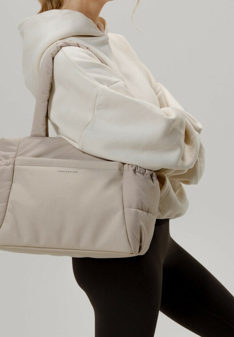 Personne portant un sweat à capuche crème et des leggings noirs, portant un grand sac fourre-tout beige rembourré sur l'épaule.